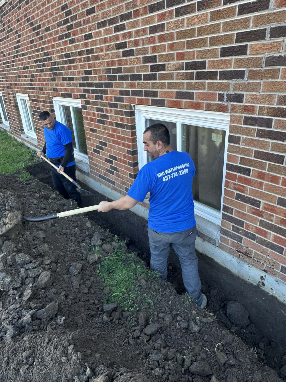 Two HRC Waterproofing crew members in branded shirts digging a trench along a foundation wall